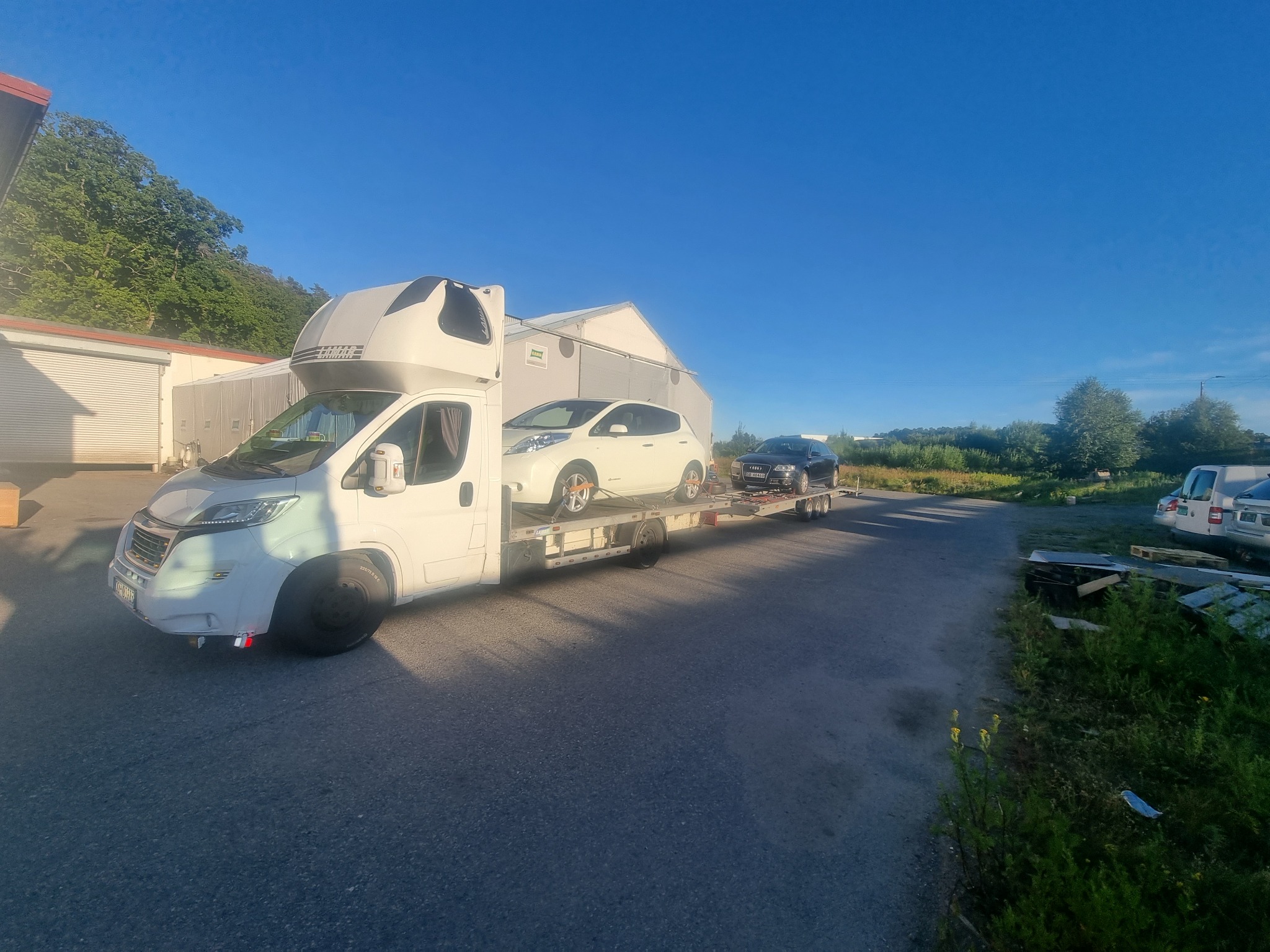 Biała laweta z zabudową kempingową przewozi dwa samochody osobowe: biały Nissan Leaf i ciemny Audi na przyczepie, na tle budynków gospodarczych i zieleni.