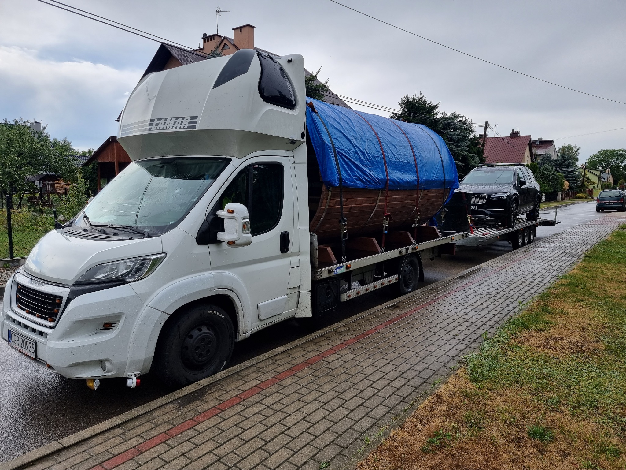 Biała laweta z przyczepą przewożąca czarny samochód osobowy i element konstrukcyjny przykryty niebieską plandeką, zaparkowana na ulicy w pochmurny dzień.