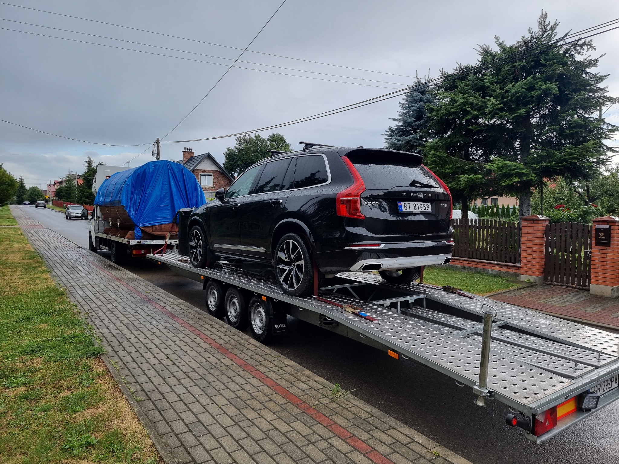 Czarny samochód Volvo XC90 na niskopodwoziowej lawecie, z widoczną tablicą rejestracyjną BT 81958, transportowany ulicą w zabudowie jednorodzinnej; w tle przykryty plandeką ładunek na drugiej lawecie.