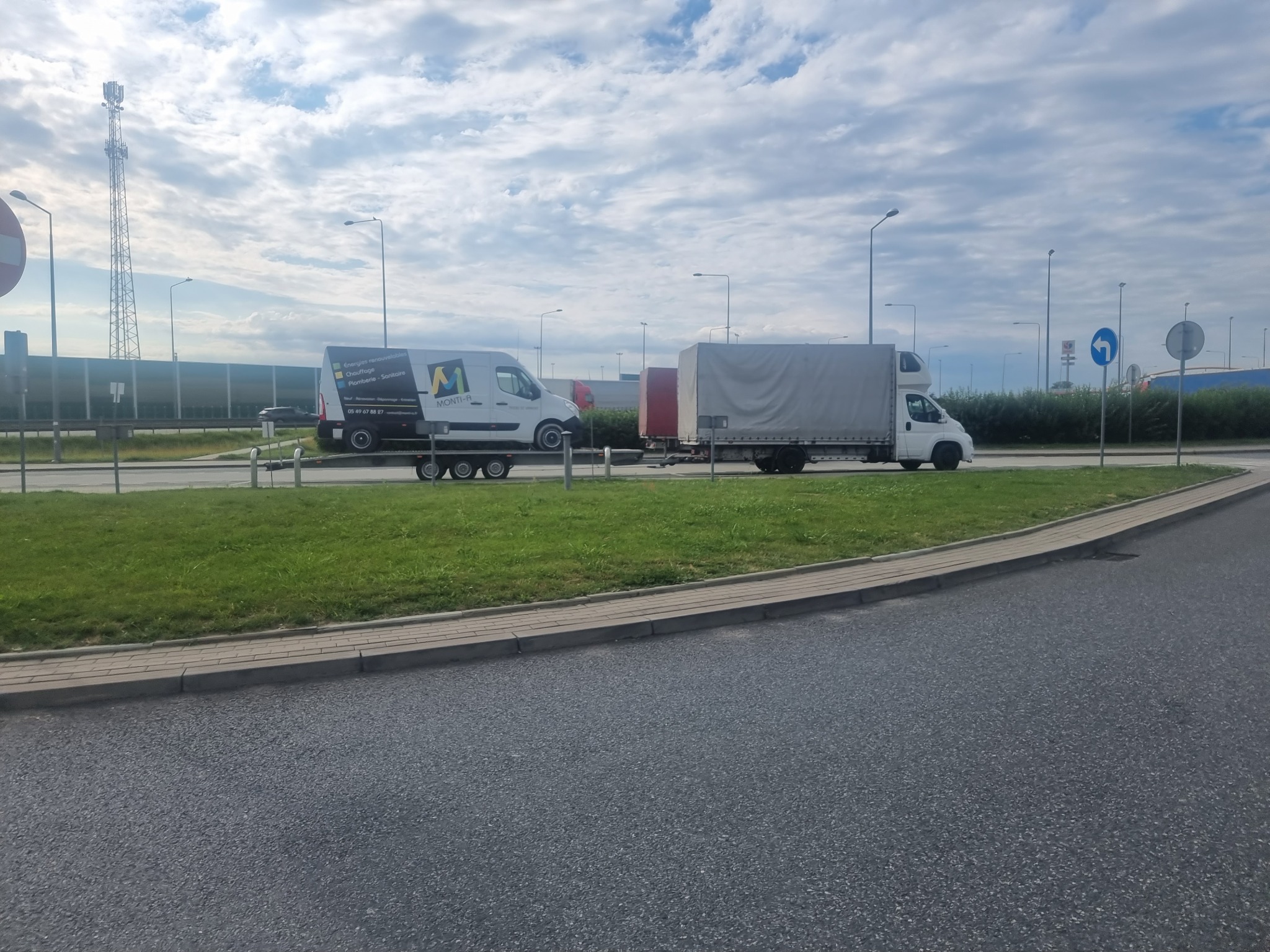 Biała laweta z przyczepą przewozi biały samochód dostawczy z logo firmy MONTI-A na tle pochmurnego nieba i infrastruktury drogowej.