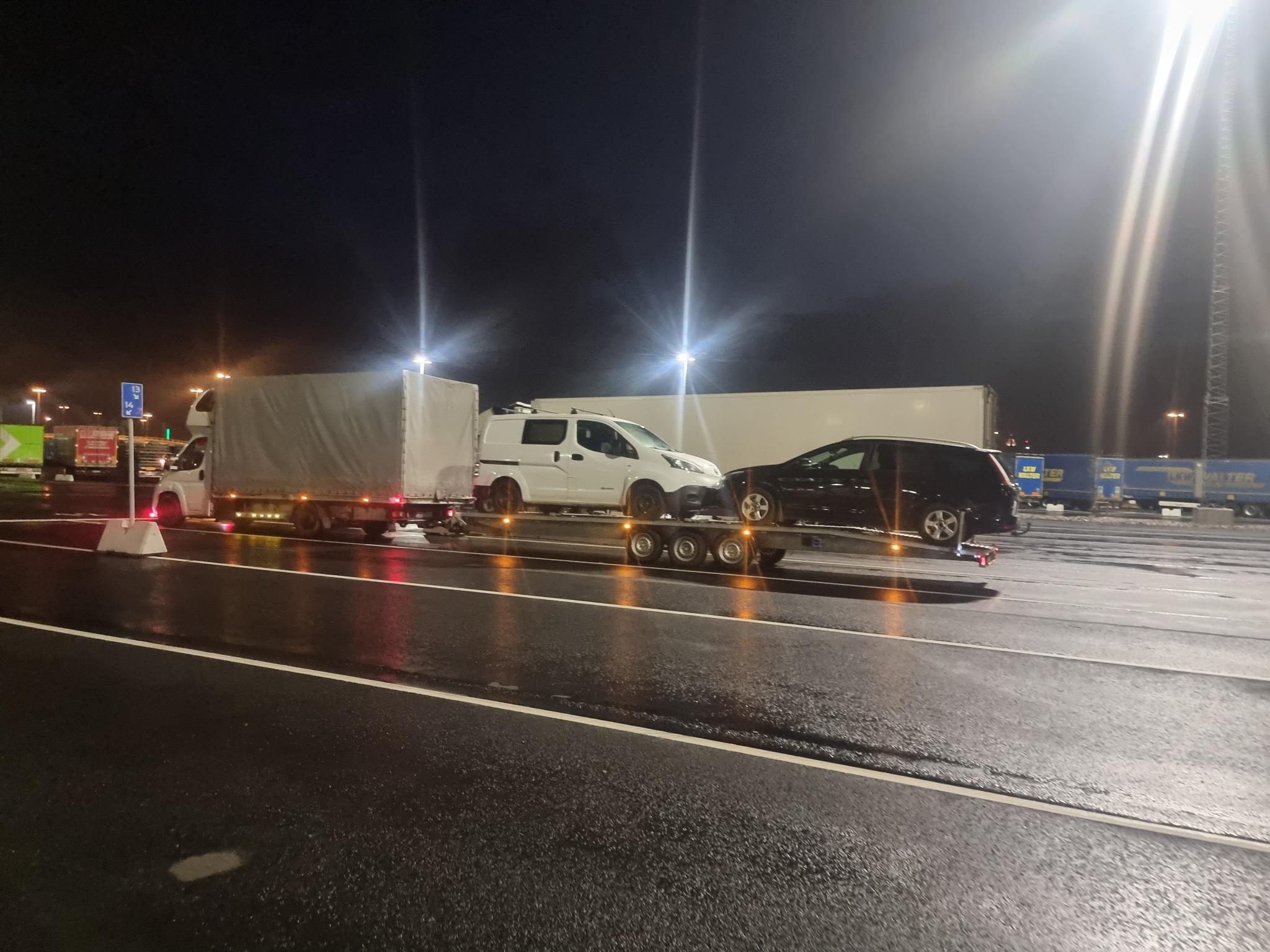 Ciężarówka z naczepą przewożąca dwa samochody: biały furgon i czarny SUV, sfotografowana nocą na mokrym asfalcie, oświetlona przez wysokie lampy.
