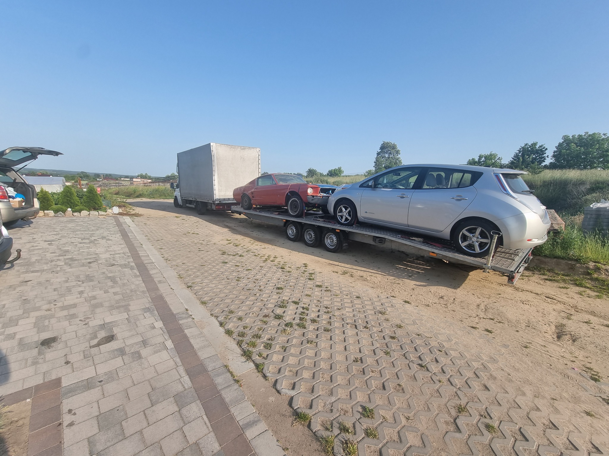 Dwa samochody, srebrny Nissan Leaf i czerwony Ford Mustang bez maski, przewożone na niskopodwoziowej przyczepie za ciężarówką na betonowym placu z kostki.