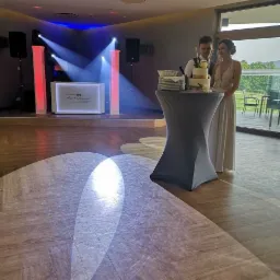 Para młoda stoi obok tortu weselnego na tle sceny z nagłośnieniem i oświetleniem, z widocznym logo DJ-a na podświetlonym panelu.