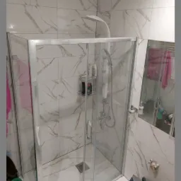 Nowoczesna kabina prysznicowa z transparentnymi drzwiami, zamontowana w łazience wykończonej białymi płytkami imitującymi marmur, widoczne półki z kosmetykami i akcesoriami.