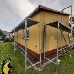 Żółty budynek jednorodzinny w trakcie czyszczenia elewacji z wykorzystaniem myjki ciśnieniowej, widoczne rusztowania i częściowo umyta ściana.