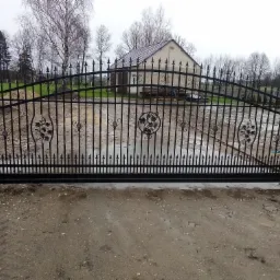 Czarna, metalowa brama wjazdowa z dekoracyjnymi elementami w kształcie winorośli, na tle szarego, pochmurnego nieba i budynku gospodarczego.