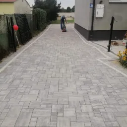 Ułożona szara kostka brukowa na podjeździe, zagęszczana mechanicznie przez pracownika, z widocznym budynkiem z numerem 39 i czarnym ogrodzeniem z czerwoną dekoracją.