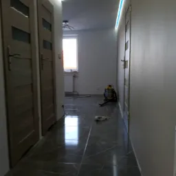 Korytarz w trakcie odświeżania: położona ciemna, błyszcząca podłoga z płytek imitujących marmur, widoczne dwie drewniane drzwi po lewej, odkurzacz przemysłowy w głębi i listwa LED pod sufitem.