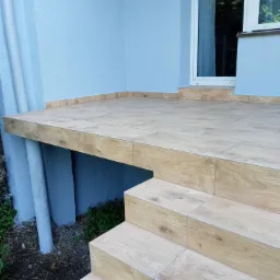 Wykończony taras z drewnopodobnych płytek, prowadzący do drzwi wejściowych, z widoczną elewacją budynku w kolorze jasnoniebieskim i rurą spustową.