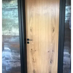 Jasne, drewniane skrzydło drzwiowe z czarną, minimalistyczną ościeżnicą i klamką na tle ściany z płytek imitujących metal.