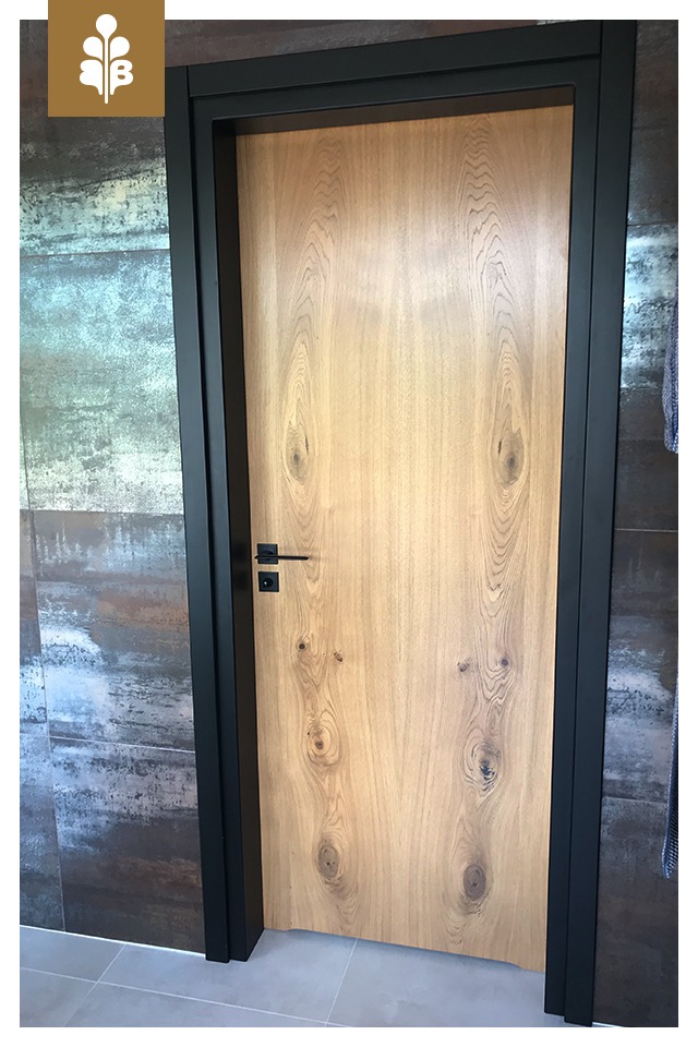 Jasne, drewniane skrzydło drzwiowe z czarną, minimalistyczną ościeżnicą i klamką na tle ściany z płytek imitujących metal.