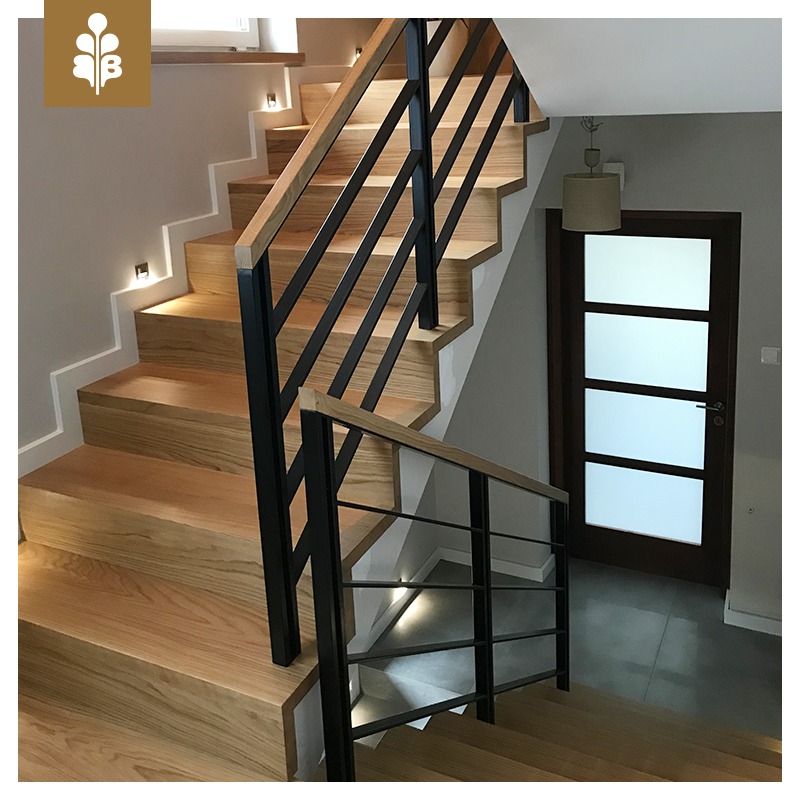 Drewniane schody z czarną metalową balustradą i oświetleniem punktowym w ścianie, prowadzące do drzwi z matowego szkła.