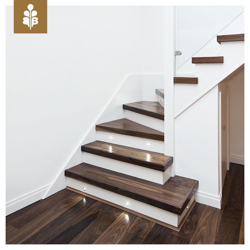 Nowoczesne schody z ciemnego drewna z białymi podstopnicami i subtelnym oświetleniem LED, widoczne na tle białej ściany i podłogi z tego samego drewna, co stopnie.