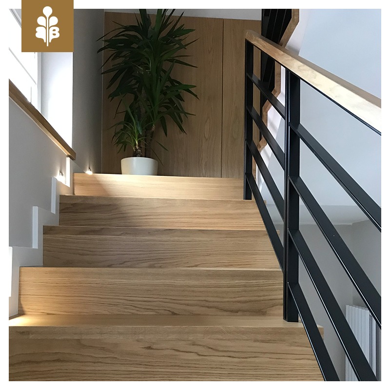 Jasne drewniane schody z czarną metalową balustradą, oświetleniem punktowym w ścianie i rośliną doniczkową na górnym stopniu.