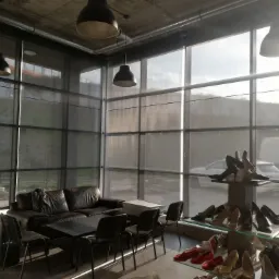 Wnętrze sklepu z dużymi oknami osłoniętymi szarymi przesłonami, widoczny skórzany narożnik i ekspozycja butów na tle ulicy.