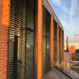 Elewacja budynku z cegły z zamontowanymi zewnętrznymi żaluzjami poziomymi, widok z balkonu na panoramę miasta w słoneczny dzień.