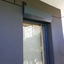 Szara kaseta zewnętrzna z widocznymi lamelami w oknie budynku mieszkalnego, z odbiciem sąsiedniego budynku w szybie.