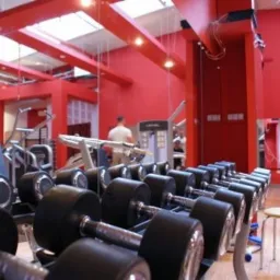 Wnętrze siłowni z rzędem hantli, czerwonymi ścianami i widocznymi maszynami do ćwiczeń w tle.