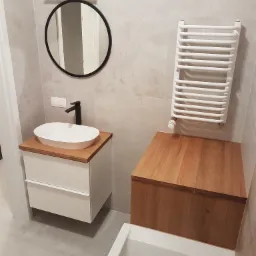 Nowoczesna łazienka w Nadarzynie z owalną umywalką na białej szafce z drewnianym blatem, okrągłe lustro z czarną ramą i grzejnik drabinkowy na ścianie z szarych płytek.