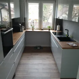 Wąska kuchnia w odcieniach szarości z drewnianym blatem, widok na okno z roślinami i drewnianą podłogą imitującą panele.