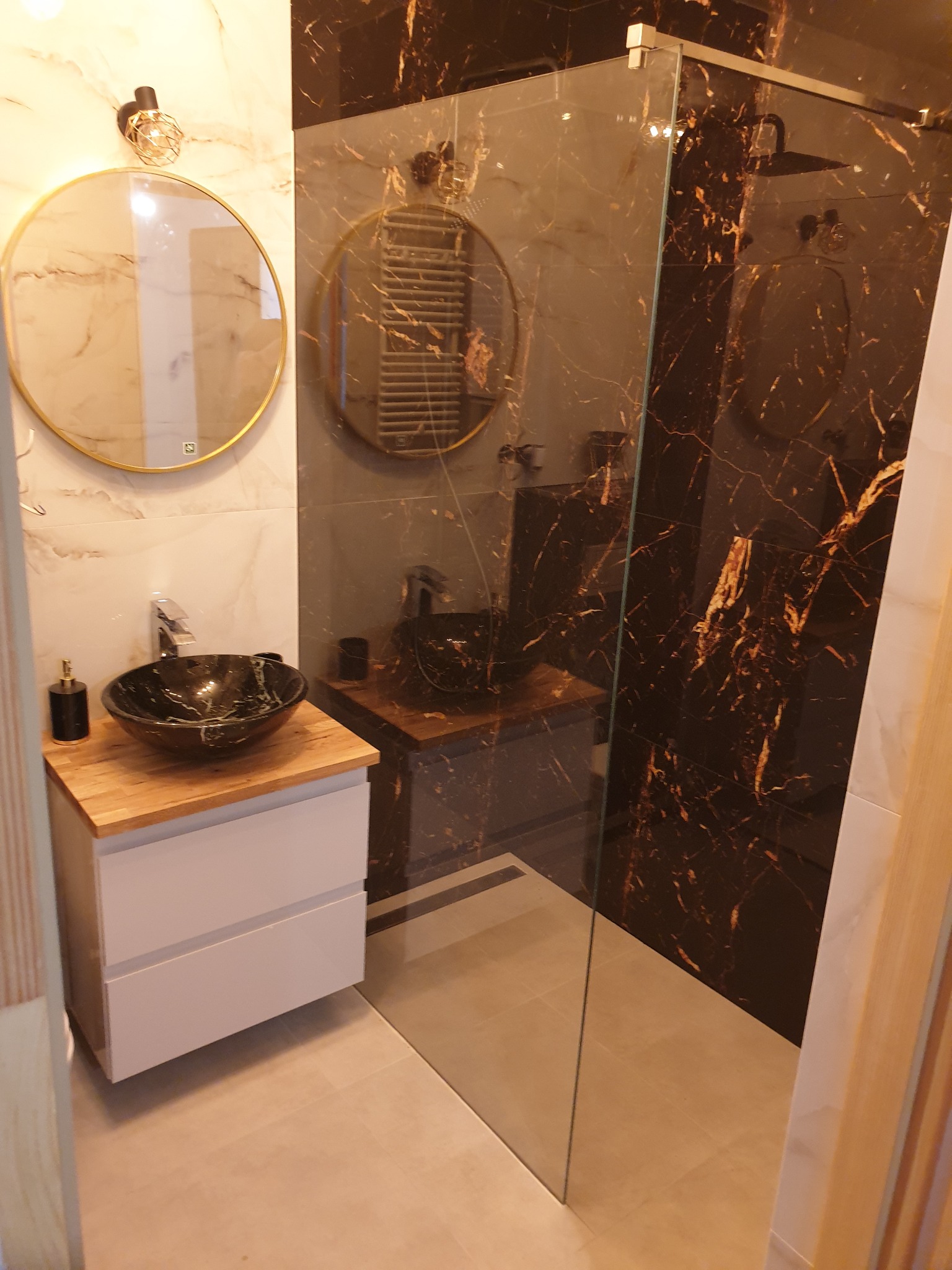 Nowoczesna łazienka w Nadarzynie z umywalką nablatową z czarnego marmuru, okrągłym lustrem z podświetleniem LED i kabiną prysznicową typu walk-in z ciemnymi płytkami imitującymi marmur.