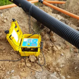 Żółta skrzynka z aparaturą pomiarową leżąca na ziemi obok czarnej rury karbowanej i pomarańczowych rur instalacyjnych, widoczne elementy łączeń.