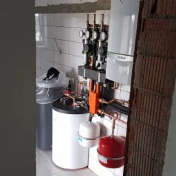 Węzeł cieplny z kotłem gazowym Vaillant, zbiornikiem buforowym i osprzętem, widoczny przez fragment surowej ściany z cegły dziurawki, w tle białe płytki i czapka Makita na śmietniku.