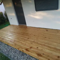 Nowo wybudowany taras z desek drewnianych przy białym budynku z czarnymi drzwiami i roletami, widok z poziomu gruntu na kamieniste podłoże.