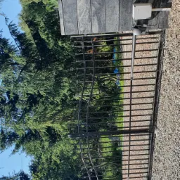 Czarna, metalowa brama automatyczna z dekoracyjnym wzorem, zamontowana na słupkach z szarego kamienia, z widocznym modułem sterującym i lampą ostrzegawczą, na tle zieleni i błękitnego nieba.