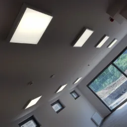 Wnętrze z zamontowanymi panelami LED w suficie i oknami z widokiem na zewnątrz, sfotografowane pod kątem, ukazujące rozmieszczenie oświetlenia i naturalne światło w pomieszczeniu.