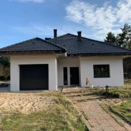 Nowoczesny, parterowy dom z czarną dachówką i białymi ścianami, widoczny z perspektywy ścieżki z czerwonej kostki, z elementami placu budowy w tle.