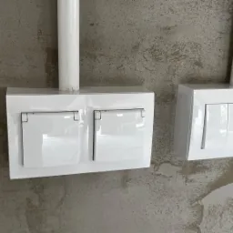 Dwa białe gniazdka elektryczne i włączniki światła zamontowane na surowej, szarej ścianie z widocznymi rurami instalacyjnymi.