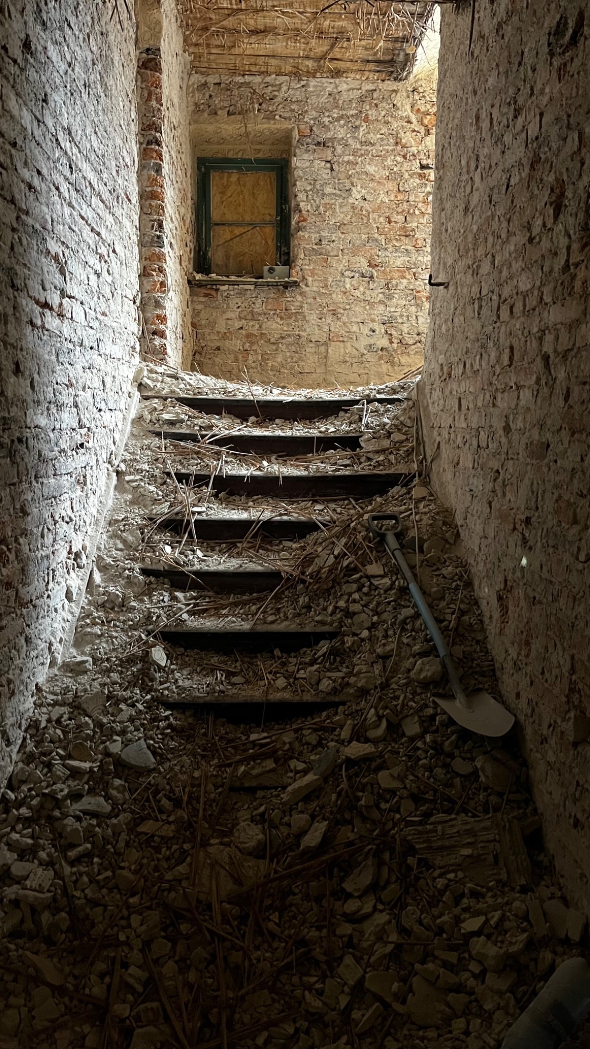 Wnętrze budynku w trakcie remontu: schody zawalone gruzem i odpadami budowlanymi, ceglane ściany z oknem zabitym deskami, widoczna łopata.