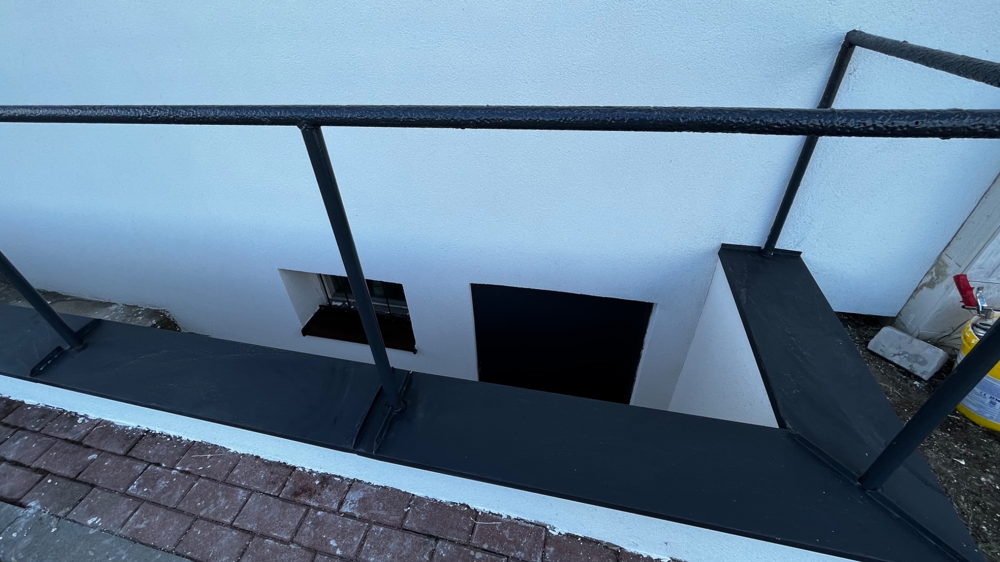 Świeżo otynkowana, biała ściana piwnicy z czarną, metalową barierką zabezpieczającą wejście, widoczna z góry, z fragmentem czerwonej ceglanej ścieżki na pierwszym planie.