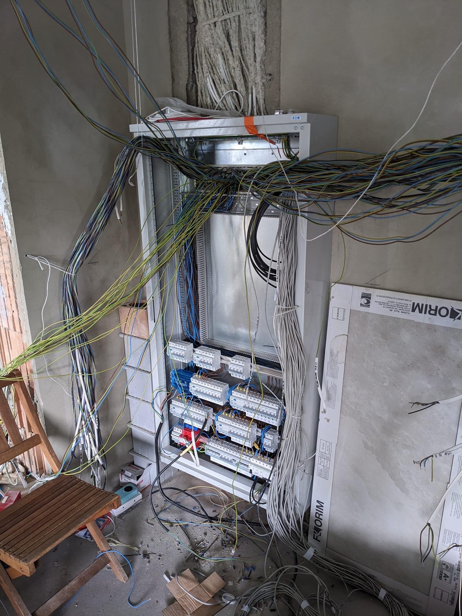 Rozdzielnia elektryczna w trakcie montażu w surowym wnętrzu, widoczne liczne kable w różnych kolorach i przekrojach, częściowo podłączone do zabezpieczeń, na podłodze resztki izolacji i narzędzia...