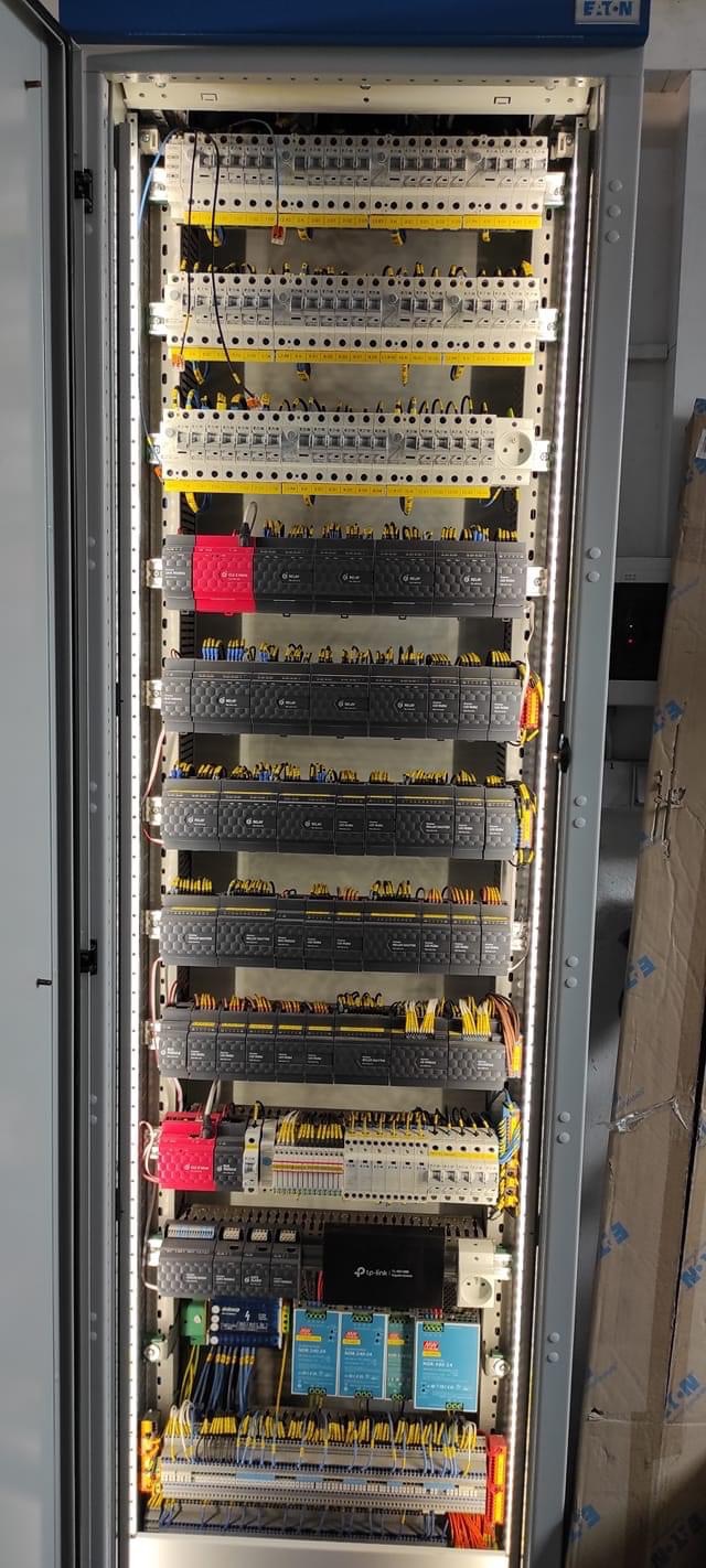 Otwarta szafa sterownicza z modułami, przewodami i zasilaczami, oświetlona listwami LED po bokach, widoczne komponenty Eaton i TP-Link.