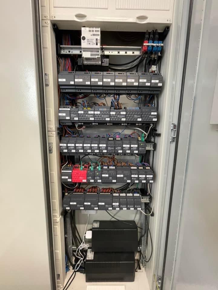 Otwarta szafa sterownicza z modułami elektronicznymi, okablowaniem i zasilaczami UPS na dole, widok z bliska na uporządkowany układ komponentów.