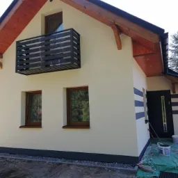 Jasna elewacja domu jednorodzinnego z drewnianymi elementami wykończeniowymi i czarnym balkonem, widoczne ślady po malowaniu i farby na ziemi.