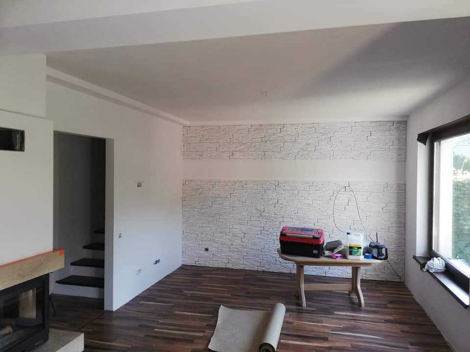 Pokój po remoncie z nową tapetą imitującą kamień na ścianie, drewnianą podłogą i stołem z narzędziami, widoczny kominek i schody w głębi