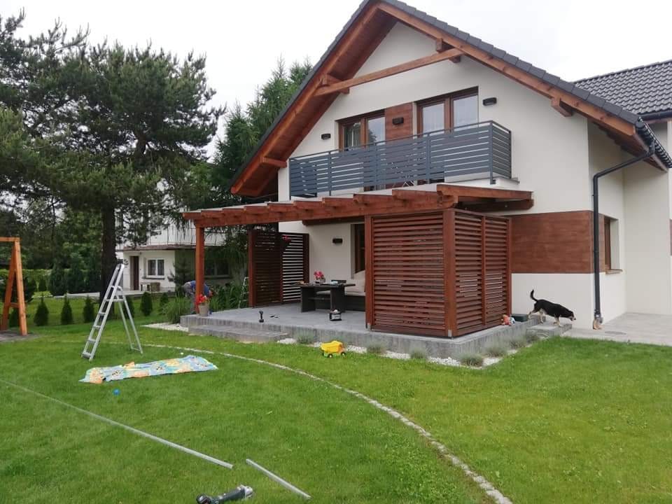 Nowoczesny dom z beżową elewacją, drewnianą pergolą i ażurowymi panelami osłaniającymi taras, widok na zadbany trawnik i elementy placu zabaw.