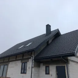 Nowy dom w trakcie budowy z ciemnoszarą dachówką, widoczne okna dachowe i orynnowanie w tym samym kolorze, ściany z białego bloczka.