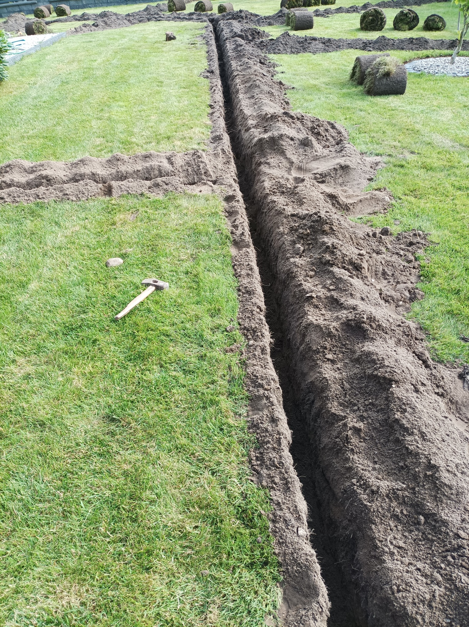 Przygotowanie terenu pod trawnik z rolki: wykopany rów, obok rolki trawy i młotek na trawie.