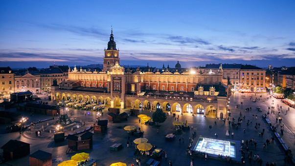 Widok z góry na Rynek Główny w Krakowie o zmierzchu, z Sukiennicami i licznymi przechodniami oraz straganami z żółtymi parasolami.