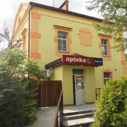 Dwupiętrowy budynek apteki w kolorze żółtym z pomarańczowymi akcentami elewacyjnymi, widoczny z zewnątrz, z wejściem zadaszonym szyldem i schodami.