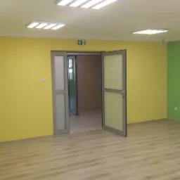 Pomalowane na żółto i zielono ściany w pustym pomieszczeniu biurowym z drewnianą podłogą, oświetlone przez panele LED w suficie, z otwartymi szarymi drzwiami prowadzącymi do innego pomieszczenia.