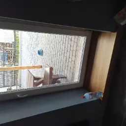 Widok przez otwarte okno na brukowany dziedziniec z drewnianą konstrukcją i elementami ogrodzenia, butelka wody i opakowanie po jedzeniu na parapecie.