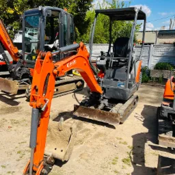 Pomarańczowa mini koparka Kubota z łyżką na placu budowy, obok inne maszyny budowlane, widoczne ślady użytkowania i ziemia.