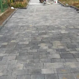 Pracownik docina szlifierką kątową szarą kostkę brukową na podjeździe, widoczna mieszarka do betonu i budynek w tle.