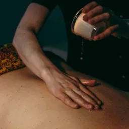 Olejowanie pleców ciepłym woskiem z naczynia podczas zabiegu relaksacyjnego; widoczny wzorzysty materiał pod ciałem.