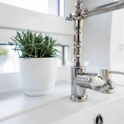 Bateria umywalkowa w stylu retro z białymi kurkami, obok doniczka z ziołami na tle okna w jasnej łazience.